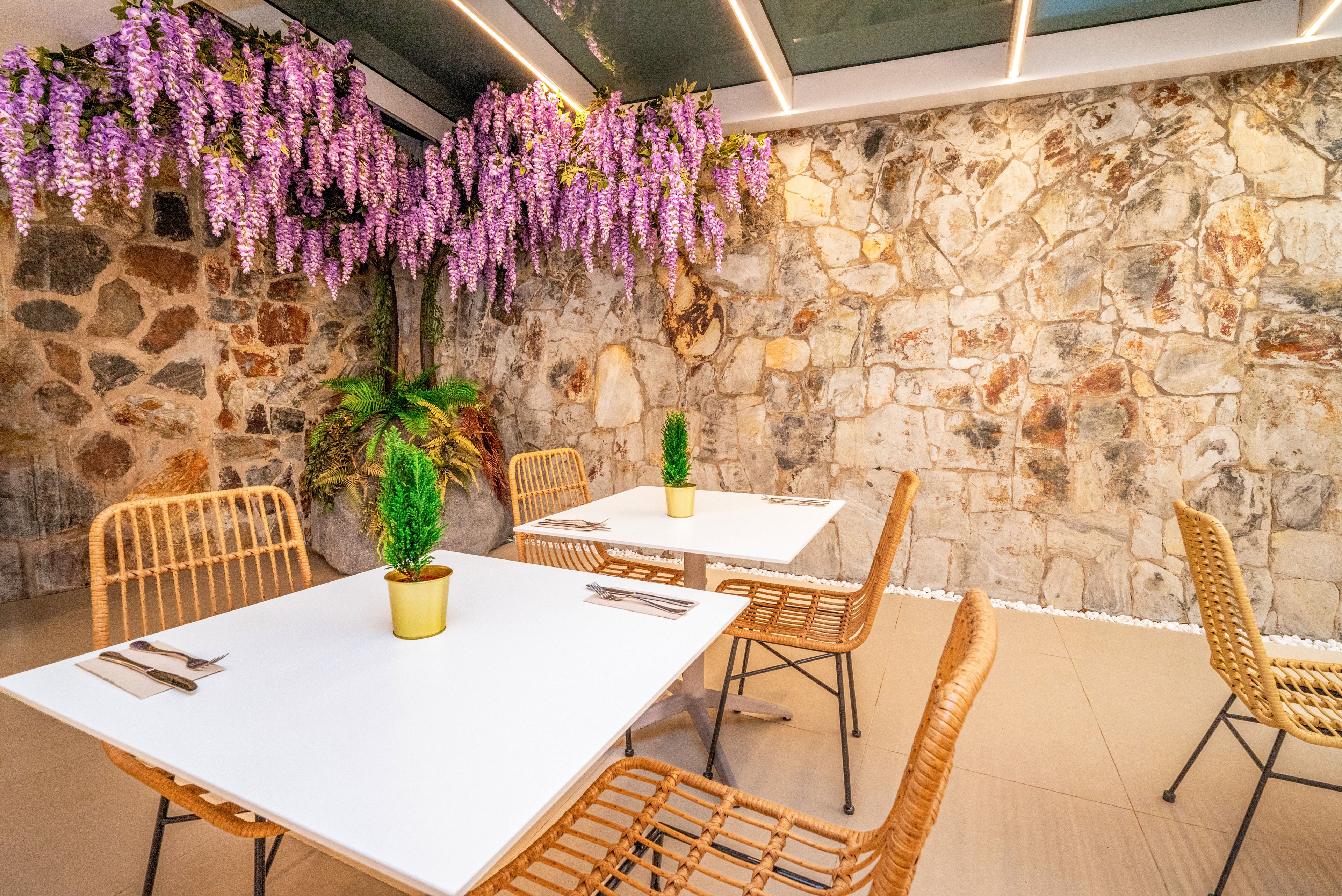 A contemporary outdoor dining area features white tables and woven rattan chairs beneath a glass ceiling. Purple hanging flowers and a lush green plant add vibrant color against a rustic stone wall backdrop. Small potted shrubs decorate each table, which is neatly set with cutlery and napkins. The space feels airy, inviting, and designed for relaxed social gatherings.