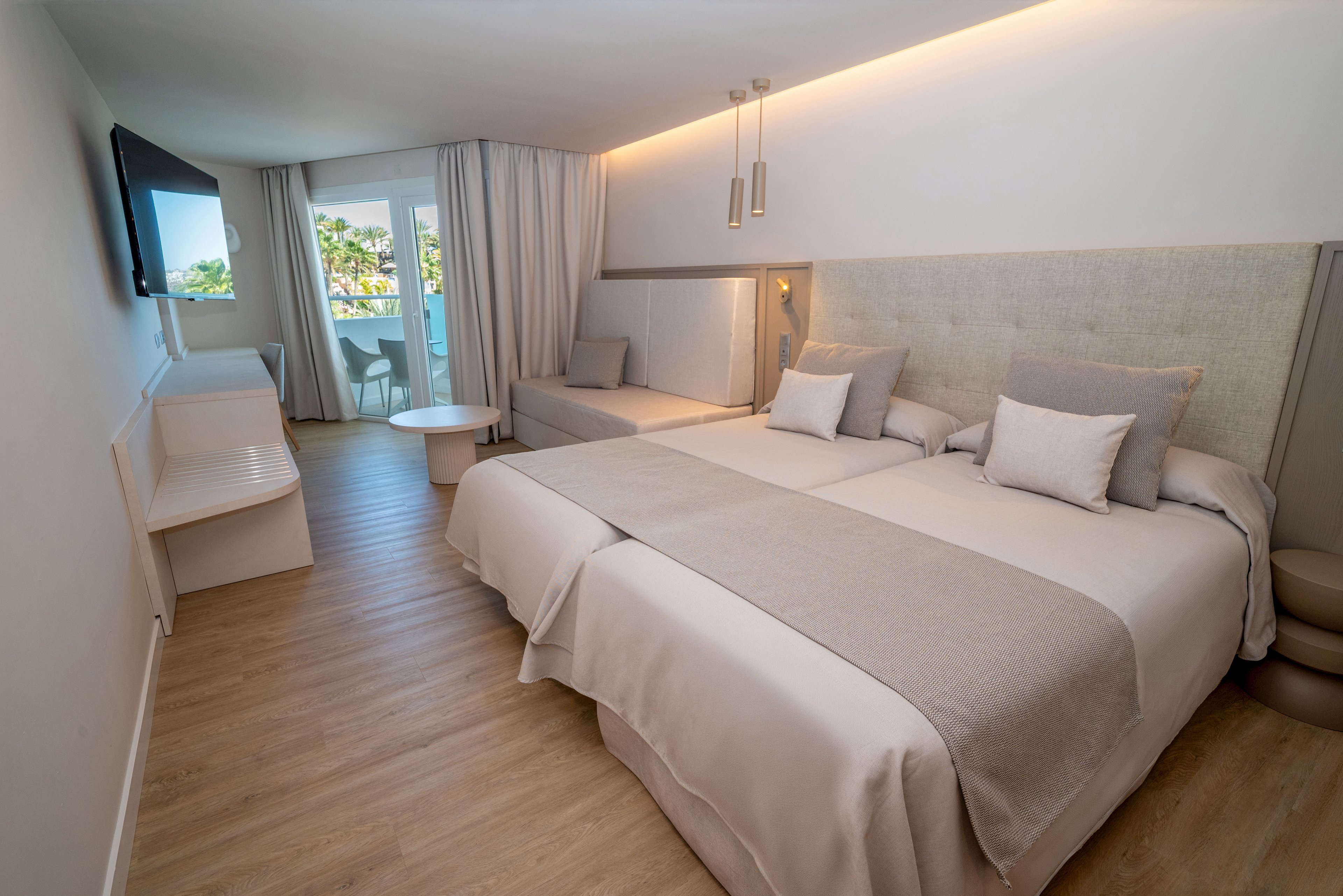 A contemporary hotel room features two large neatly made beds with neutral bedding and multiple pillows. The space includes a wall-mounted flat-screen TV, a small sofa or daybed, and a round table near the balcony. Floor-to-ceiling curtains frame the glass doors that open to a balcony with a view of greenery and palm trees. Soft, indirect lighting and light wood flooring create a calm, minimalist atmosphere.