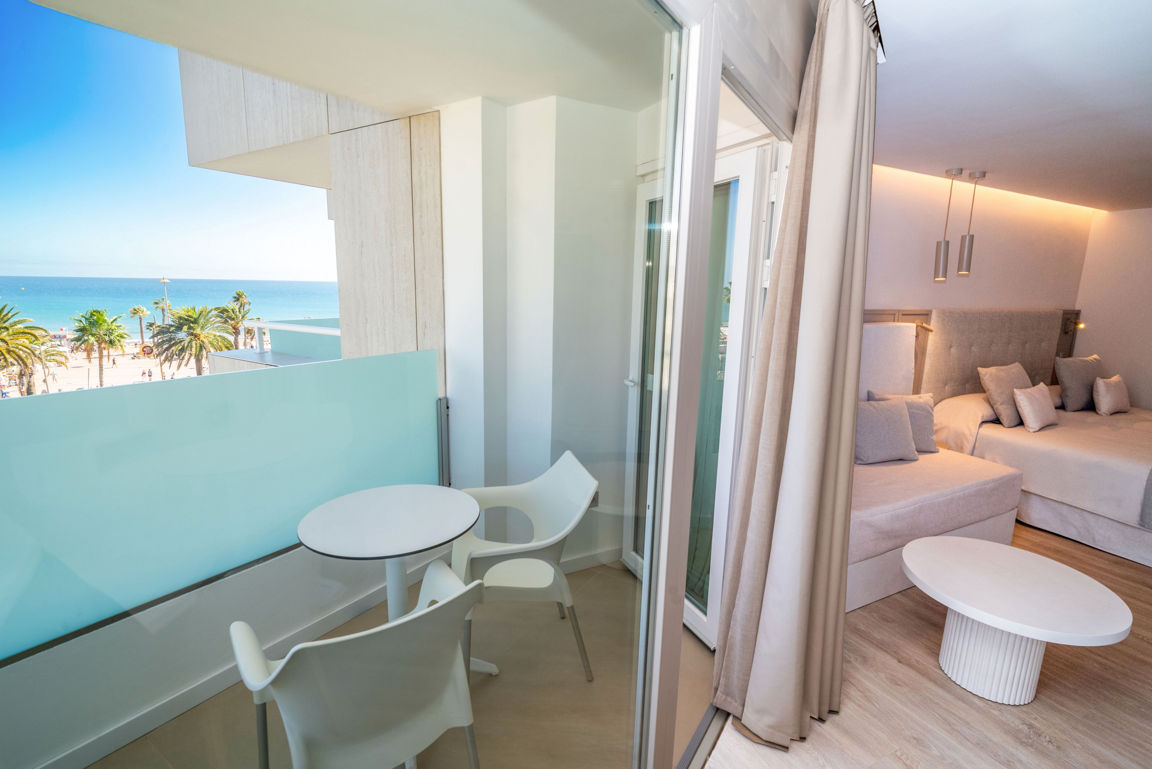 Bright contemporary hotel room opens onto a small balcony overlooking a sunny beach and ocean. Inside, a large bed with plush pillows and soft lighting creates a cozy, minimalist atmosphere. The balcony features a round white table and two chairs, framed by glass panels and sliding doors. Palm trees, blue sky, and calm sea complete the relaxing coastal setting.