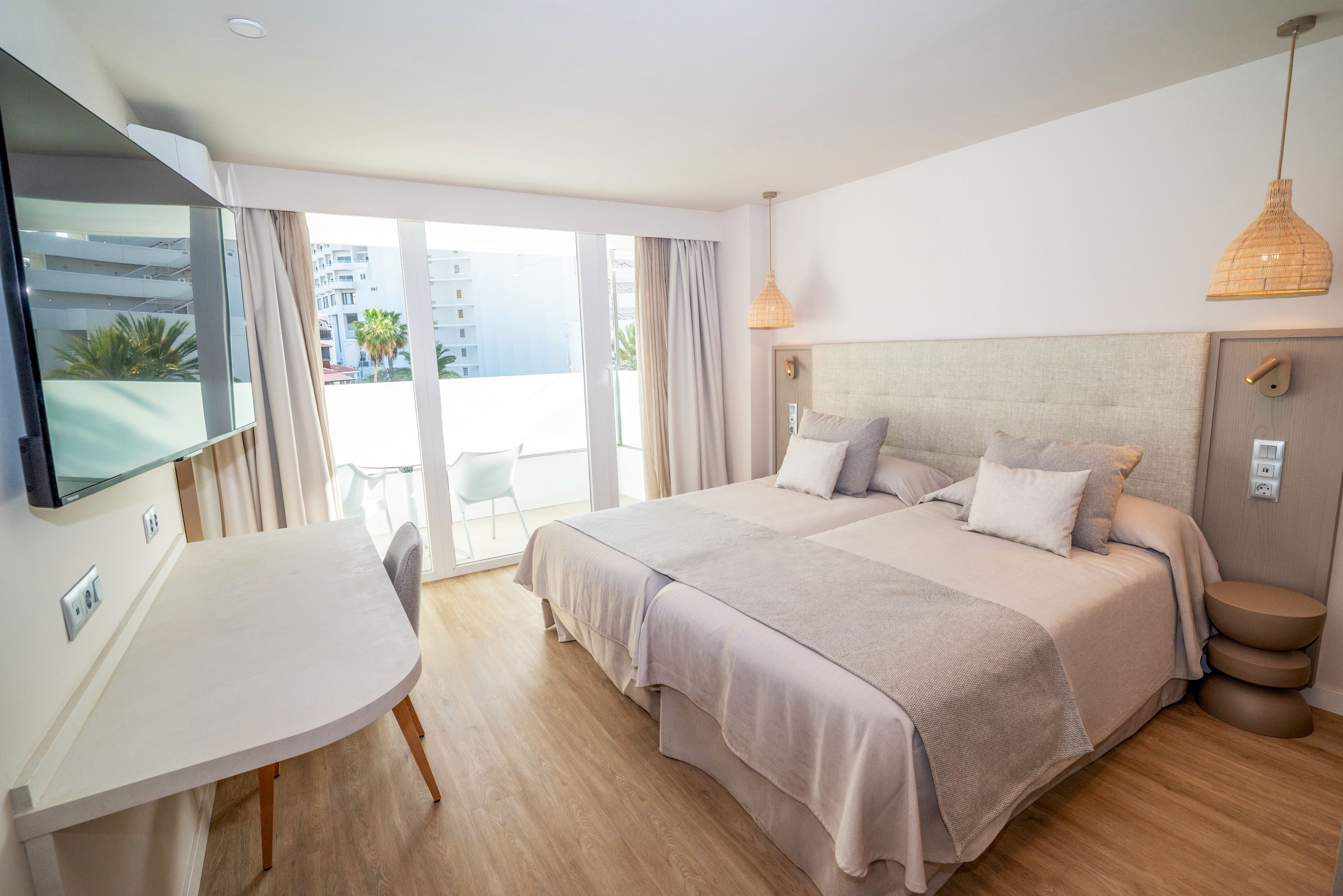 Contemporary hotel room featuring two neatly made twin beds with neutral bedding and cushions. A wall-mounted flat-screen TV faces a sleek white desk along the left side of the room. Large glass doors open to a balcony with outdoor chairs, allowing natural light to fill the space. Light wood flooring, woven pendant lamps, and soft beige tones create a calm, minimalist atmosphere.