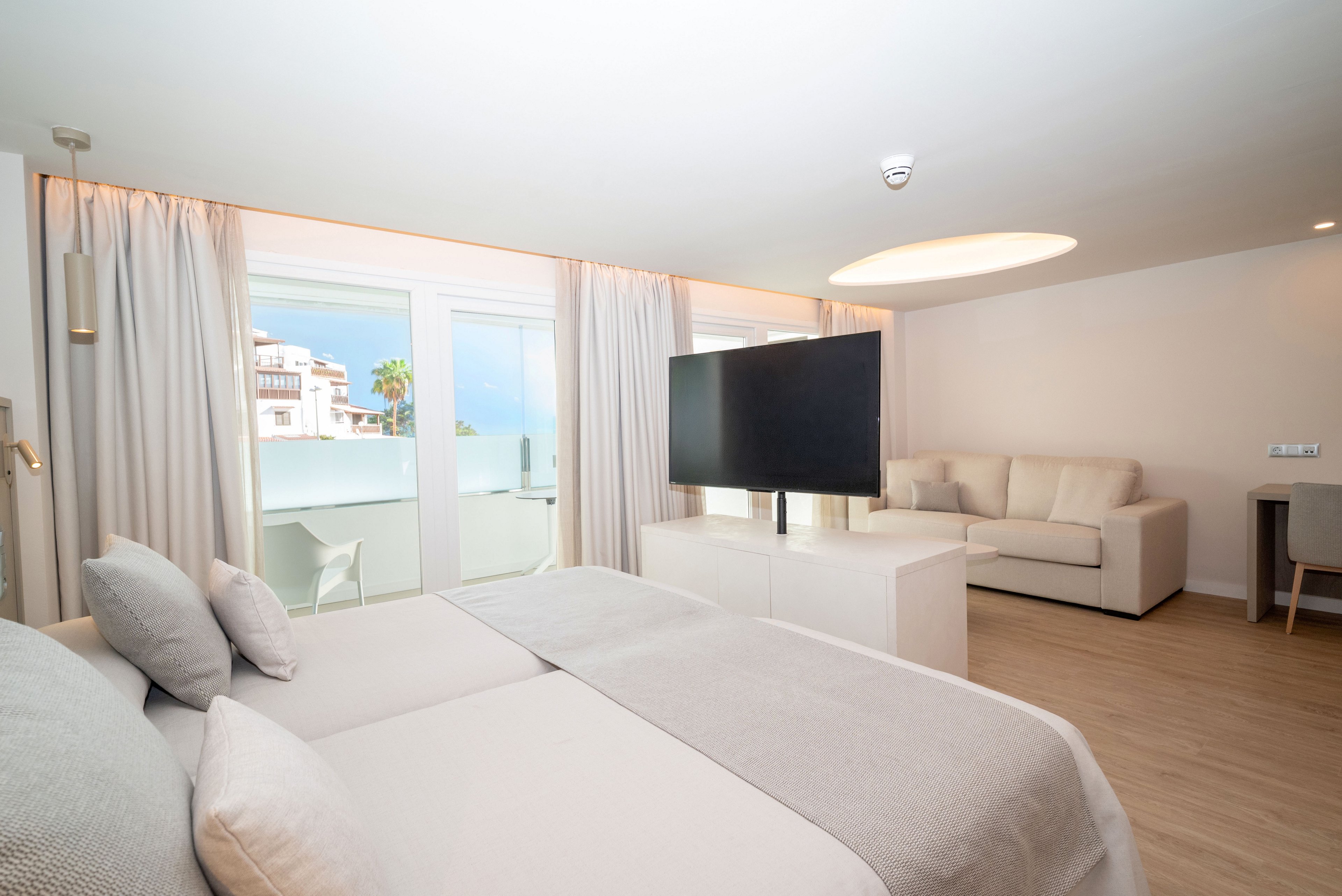 A spacious modern bedroom features a large neatly made bed facing a flat-screen television on a central stand. Floor-to-ceiling curtains frame glass doors that open to a balcony with a partial coastal view. A light-colored sofa and small desk sit against the far wall, complementing the minimalist decor. Soft neutral tones and natural light create an airy, relaxing atmosphere ideal for a hotel or vacation rental.