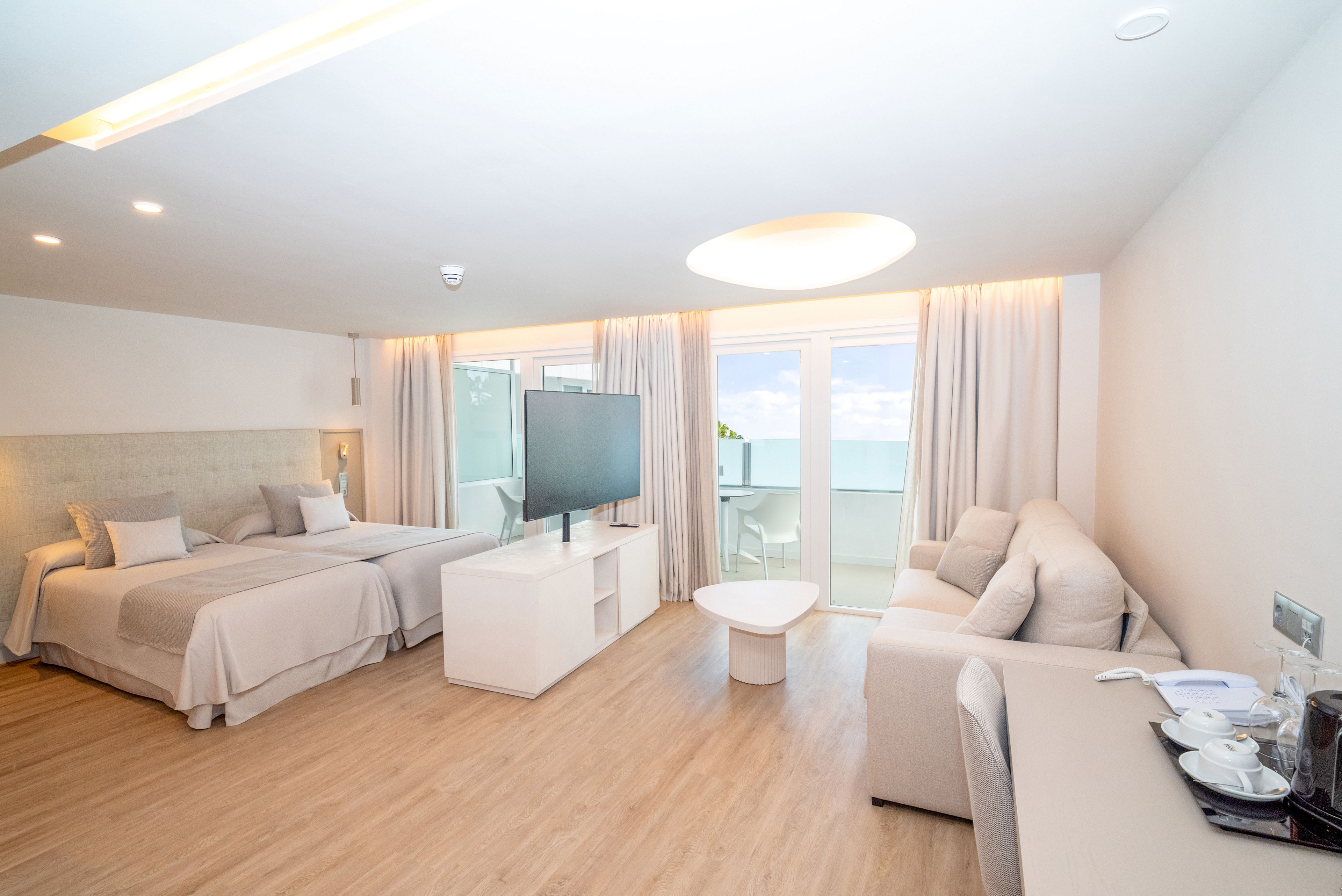 Spacious contemporary hotel room features a large bed and a sofa in a bright, open layout. A flat-screen TV stands on a central console facing the seating area. Floor-to-ceiling windows with sheer curtains open onto a balcony overlooking the ocean. Light wood flooring and white furnishings create an airy, minimalist atmosphere. A small coffee station is visible on the right side of the room.