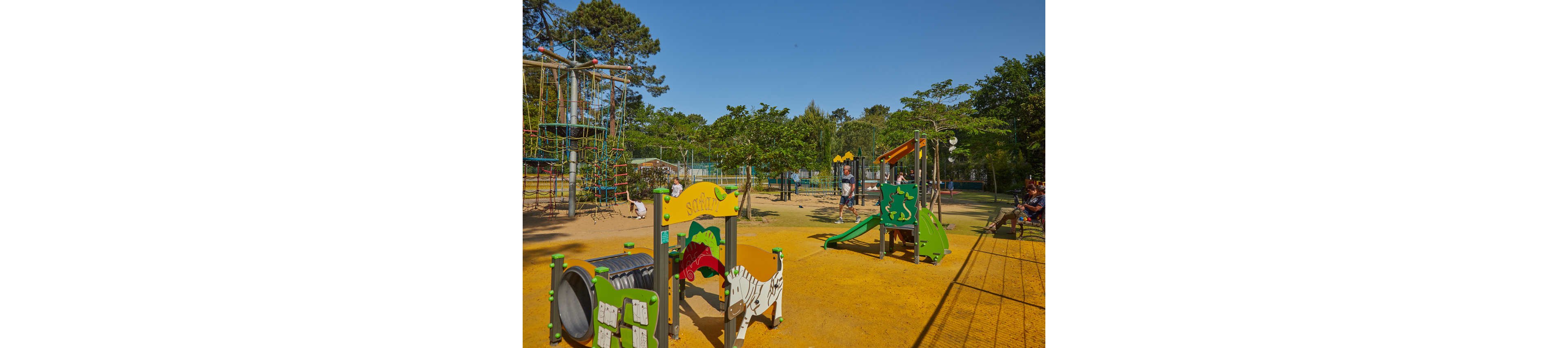 A bright outdoor playground features various colorful play structures, including slides, climbing frames, and tunnels. Children are seen playing and exploring the equipment under a clear blue sky. The ground is covered with a yellow safety surface, and trees surround the play area providing a natural backdrop. The scene conveys an active, family-friendly environment in a public park.