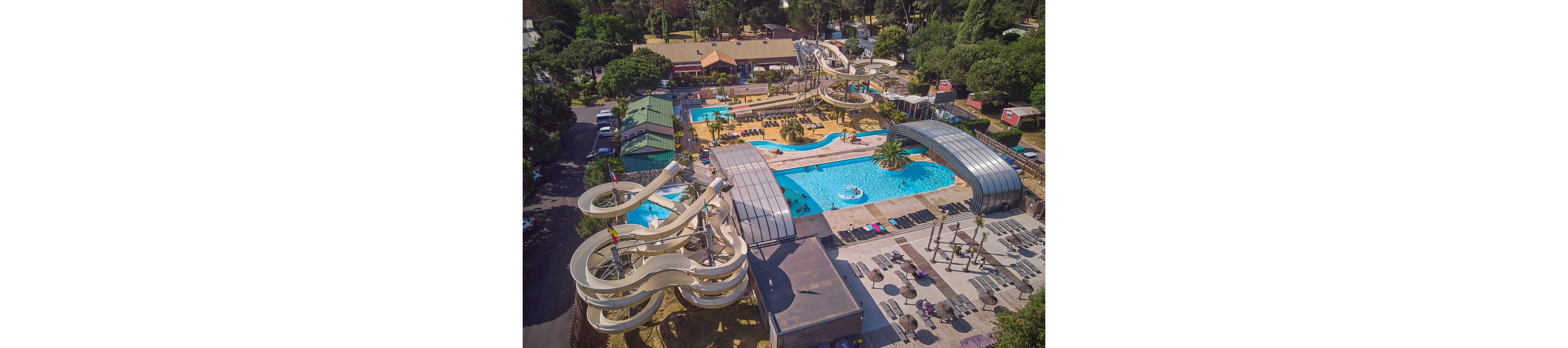 A large outdoor water park is captured from above, showcasing multiple twisting water slides and bright blue swimming pools. The complex includes sun loungers, shaded seating areas, and covered pool sections surrounded by trees. Buildings and facilities line the edges of the park, indicating amenities for visitors. The overall scene suggests a busy recreational destination designed for family fun and summer relaxation.