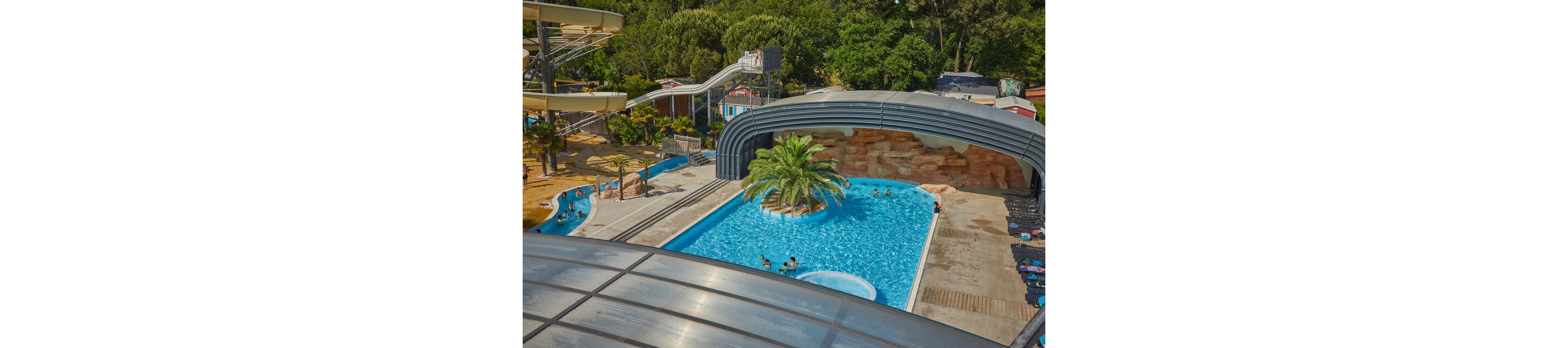 An outdoor water park features a bright blue swimming pool surrounded by palm trees and greenery. Several people relax and play in the water near a central palm tree island. Curved water slides and a covered pool area create a dynamic recreational setting. The scene is captured on a sunny day, emphasizing leisure and summer vacation vibes.