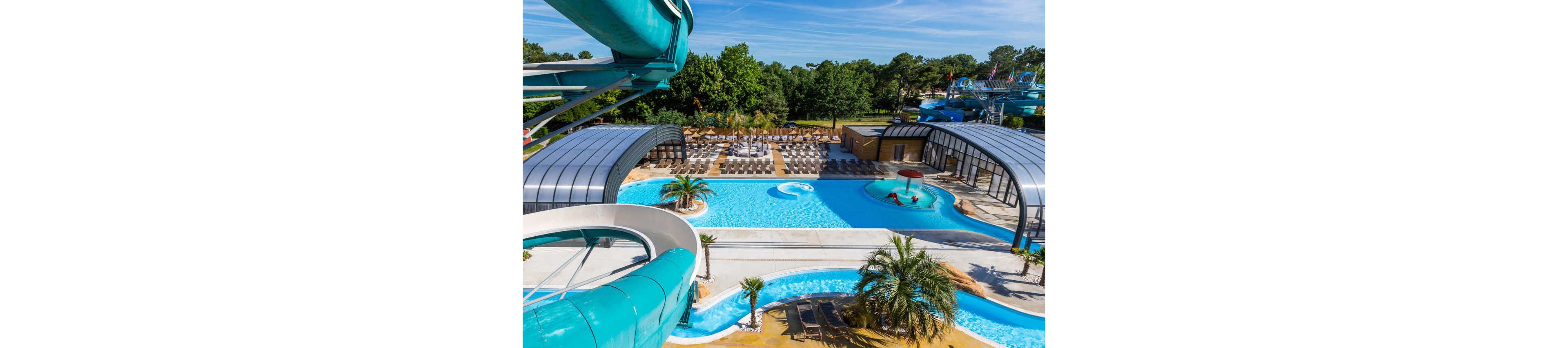 A large outdoor water park features multiple bright blue swimming pools surrounded by lounge chairs. Curved water slides frame the scene, leading into a lazy river-style channel and main pool. Two semi-enclosed pool areas with arched roofs provide shaded swimming spaces. Palm trees and greenery add a tropical resort atmosphere, while dense forest borders the park in the background.
