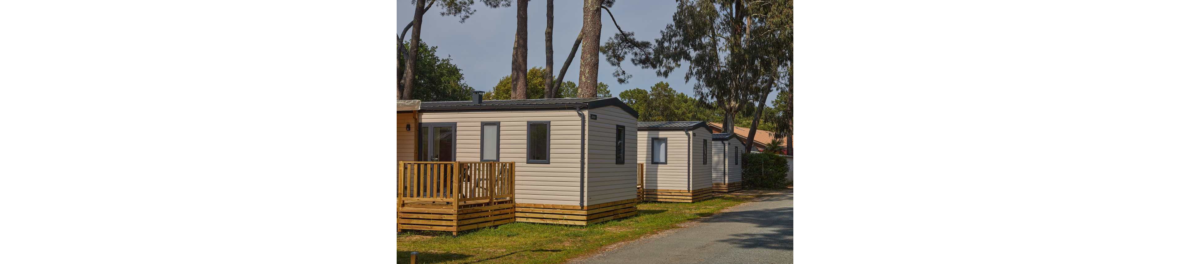 Several modern mobile homes are lined up along a quiet campsite road. Each unit features light-colored siding, dark roofs, and small wooden decks or porches. Tall trees surround the area, providing shade and a natural backdrop. The scene suggests a peaceful holiday park or vacation rental setting in a wooded environment.