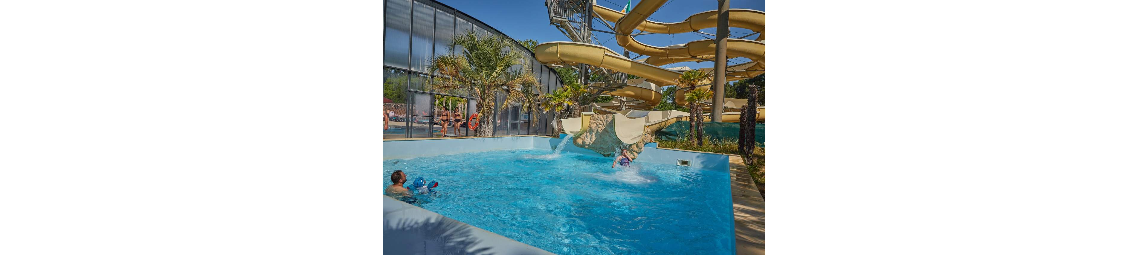 An outdoor swimming pool features a large twisting water slide emptying into the clear blue water. A child splashes out of the slide while another person watches from the shallow end with inflatable toys. Palm trees and tropical plants line the poolside, creating a resort-like atmosphere. A glass-walled building and lifebuoys are visible in the background, suggesting a family-friendly aquatic park.