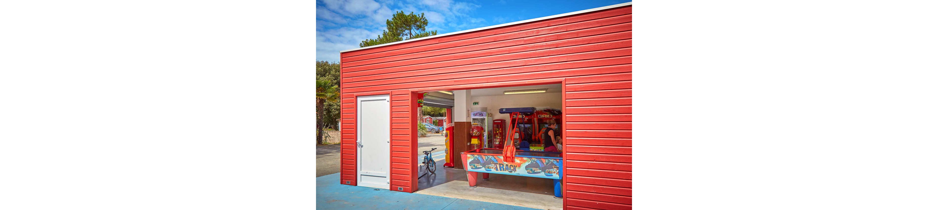 A small red-sided arcade building stands open to the outside, revealing several colorful game machines inside. A child is seated at an air hockey style game table near the entrance, surrounded by bright graphics and flashing lights. A bicycle is parked just outside on the blue pavement, suggesting a casual stop during outdoor play. Trees and other structures are visible in the background under a partly cloudy sky, giving the scene a relaxed holiday atmosphere.
