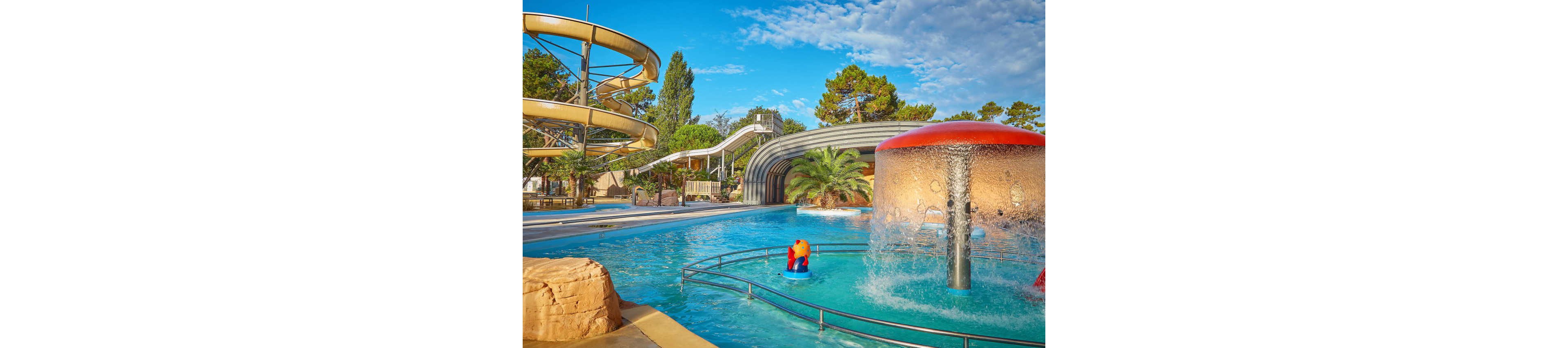 An outdoor water park features a bright blue swimming pool with a large red-topped mushroom fountain in the foreground. A spiral yellow water slide and a long straight slide curve above the pool area, surrounded by trees and greenery. A small children’s ride shaped like an animal sits in the shallow water near safety railings. The scene is sunlit under a partly cloudy blue sky, creating a fun and inviting holiday atmosphere.