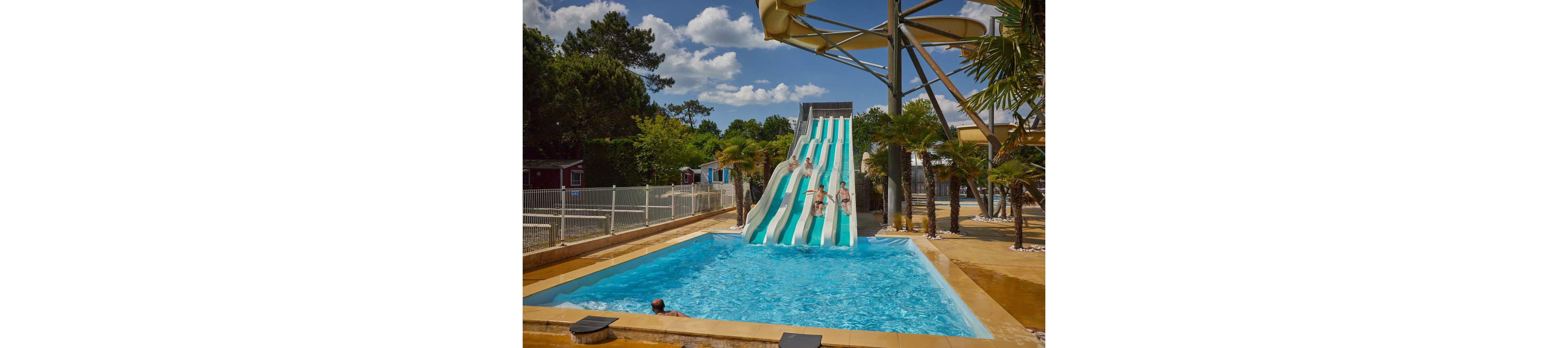 Several people slide down parallel water slides into a bright blue pool at an outdoor water park. The scene is set on a sunny day with clear skies, surrounded by trees, palm trees, and nearby cabins or small buildings. Adults and children are enjoying the ride, creating splashes as they reach the pool. A person in the foreground watches from the pool, adding to the lively, recreational atmosphere.
