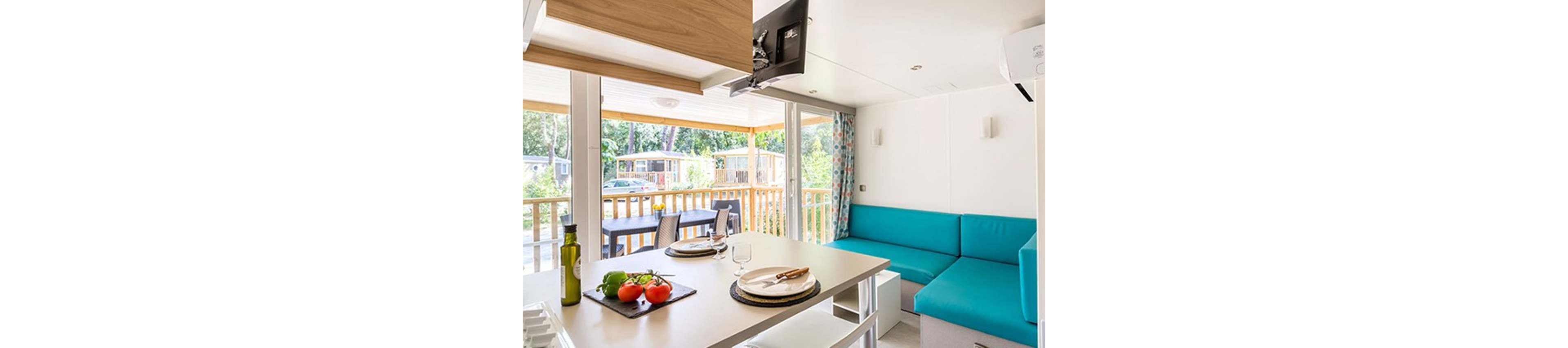 A modern mobile home kitchen opens onto a sunny wooden terrace through large sliding glass doors. Inside, a turquoise corner sofa and compact dining table create a cozy living area. The kitchen counter holds fresh vegetables, a bottle of olive oil, and neatly set plates and glasses. Outside, a table and chairs sit on the deck, with neighboring cabins and trees visible in the background.