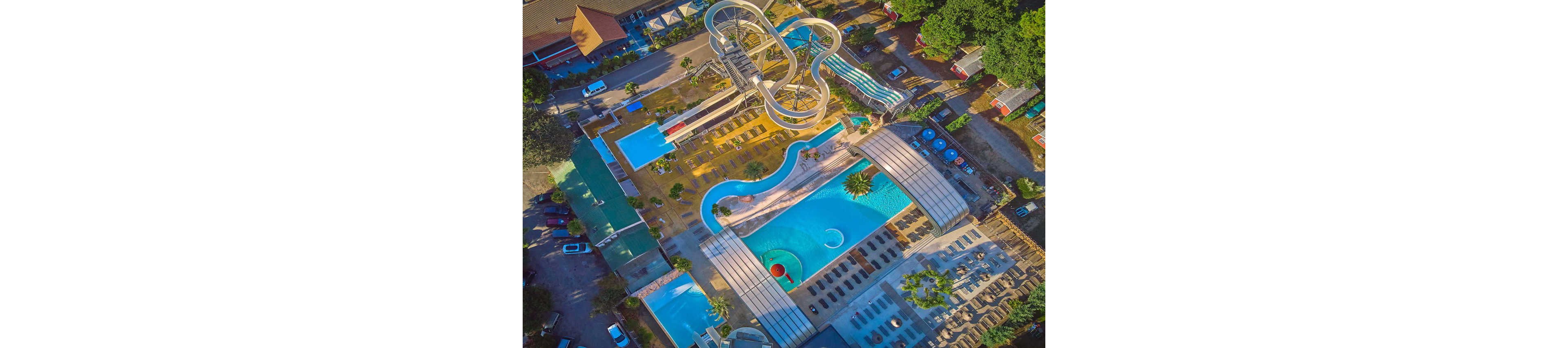 A large outdoor water park is captured from above, showcasing multiple pools and winding water slides. The complex includes a main swimming pool, smaller rectangular pools, and a lazy river-style channel. Sun loungers are neatly arranged around the pools, with shaded areas and surrounding buildings visible. Bright blue water and warm-toned walkways create a vibrant, summery atmosphere.