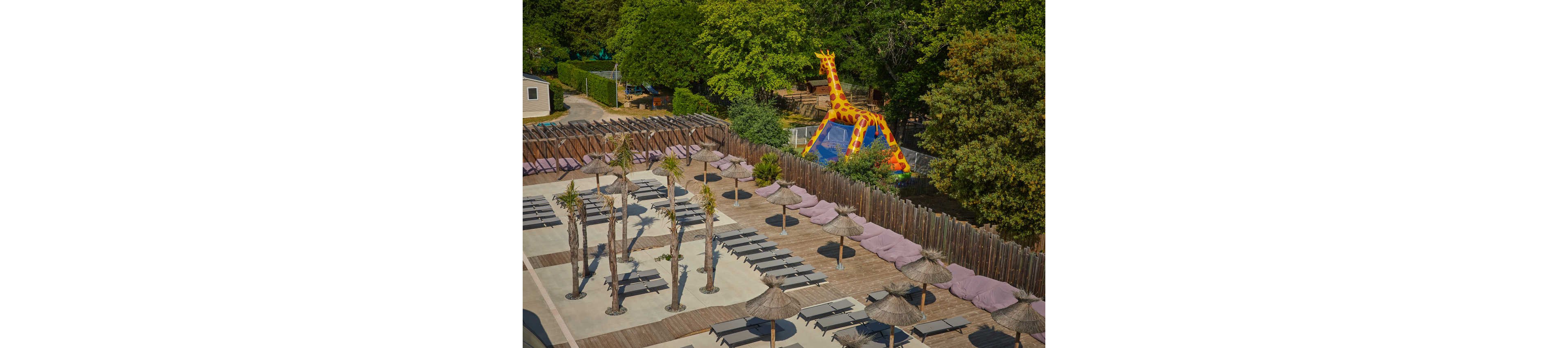 An elevated view shows an empty resort-style sun terrace lined with lounge chairs and straw parasols. Artificial palm trees and wooden decking create a tropical atmosphere within a fenced area. In the background, a large inflatable slide shaped like a giraffe stands near dense green trees. The scene appears calm and quiet, suggesting an off-peak or early-morning moment at a family-friendly leisure facility.