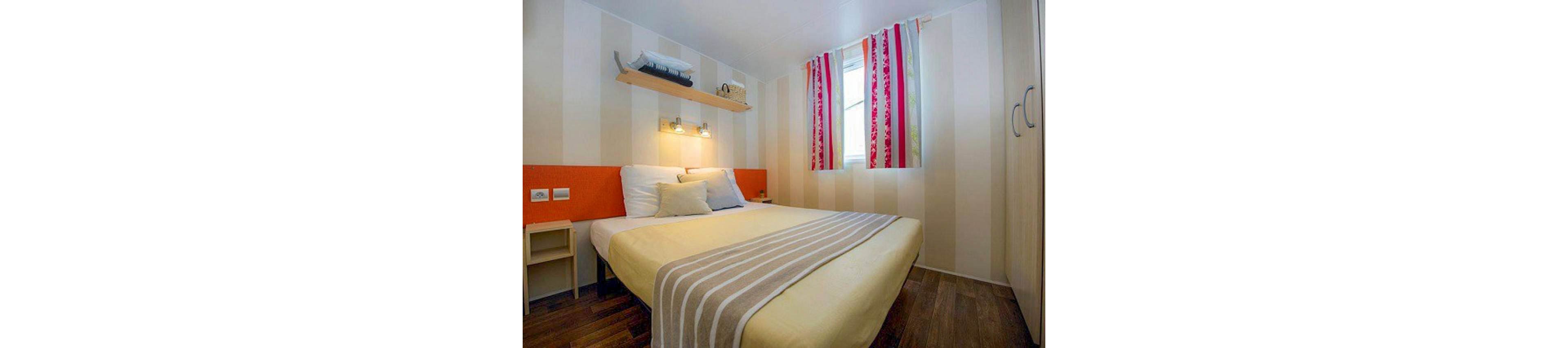 A compact modern bedroom features a neatly made double bed with yellow bedding and a striped throw. Soft wall-mounted lights illuminate the headboard and pillows, creating a warm and inviting atmosphere. A shelf above the bed holds folded towels and decor, while striped curtains add a pop of color to the window. Light-colored walls and wooden flooring enhance the cozy, contemporary feel of the space.