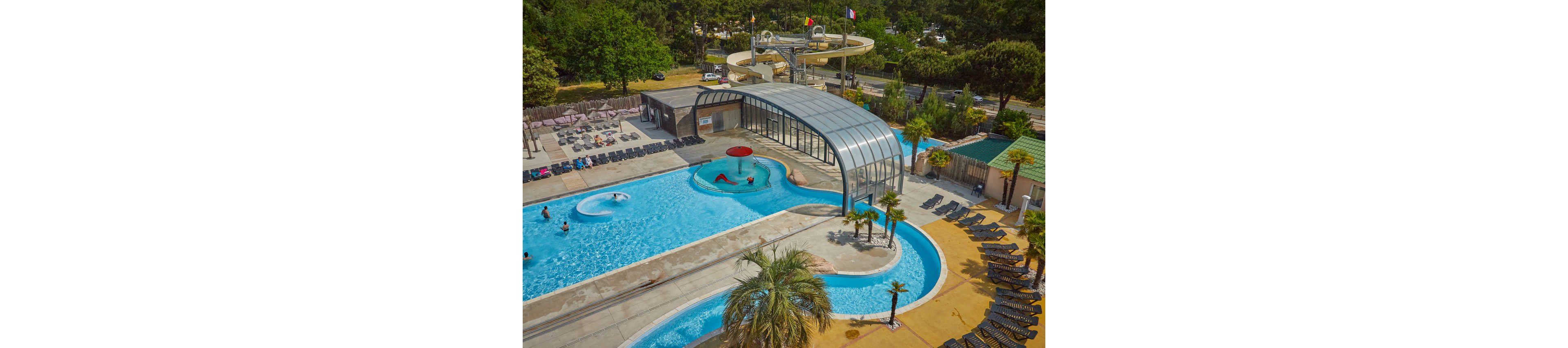 A large resort-style swimming pool complex is seen from above, featuring multiple interconnected pools and a partially covered section with a curved glass roof. Several people relax and play in the water, including a child under a red mushroom fountain. Surrounding the pools are rows of empty lounge chairs, palm trees, and a waterslide structure in the background. The setting is bright and sunny, with green trees and a nearby road visible beyond the pool area.