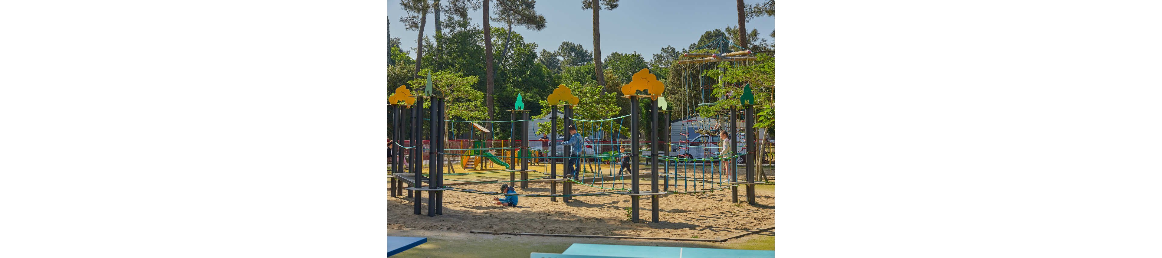 Children play on a large outdoor rope playground structure in a sandy area. The scene shows several kids climbing and balancing on nets and bridges while one child sits in the sand. Tall trees and greenery surround the play area, creating a natural park setting under a clear sky. Bright colors on the equipment and soft sunlight give the image a cheerful, active atmosphere.