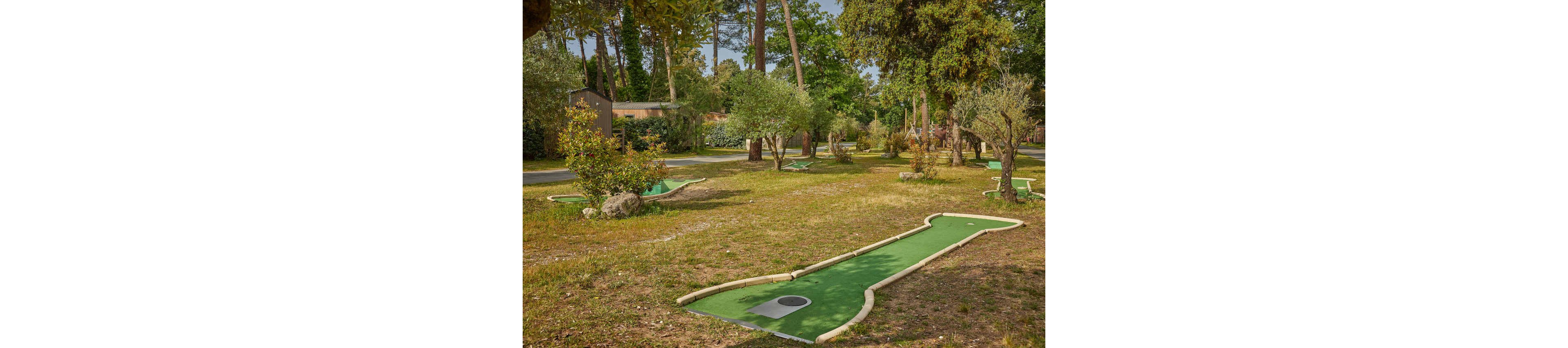An outdoor mini golf course is set within a grassy, tree-filled park. Several putting lanes with green artificial turf and light-colored borders curve through the open space. Bushes, rocks, and mature trees create a natural, shaded environment around the holes. A small building and pathway are visible in the background, suggesting a recreational or holiday setting.