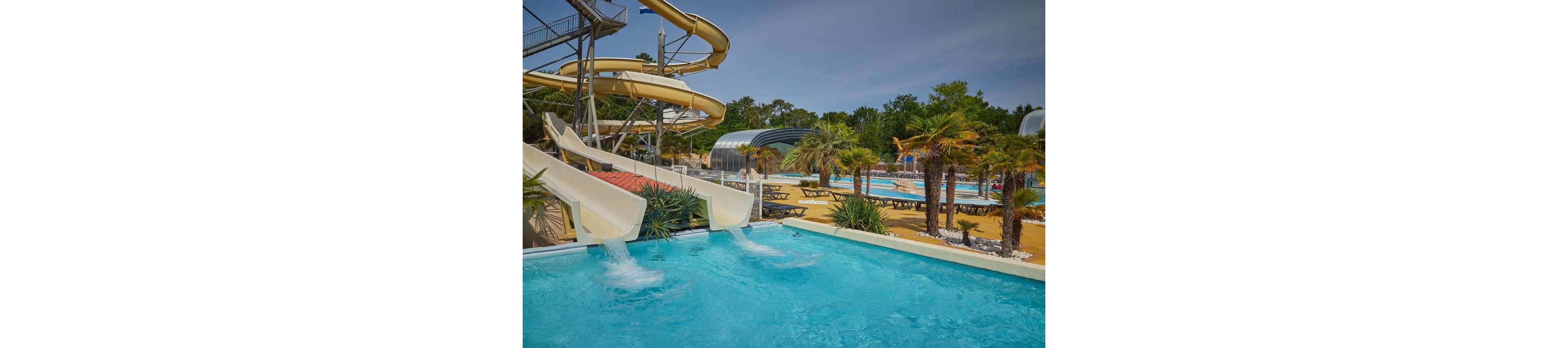 An outdoor water park features multiple curving water slides emptying into a bright blue pool. In the background, additional swimming pools and a covered pool area are visible, surrounded by lounge chairs and picnic tables. Palm trees and tropical plants line the pool deck, creating a resort-like atmosphere. The scene is sunlit under a clear sky, suggesting a warm summer day ideal for family recreation.