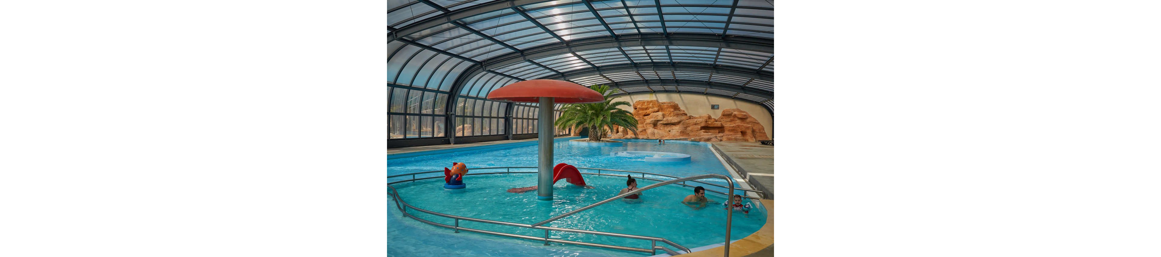 An indoor swimming pool features a large red mushroom-shaped fountain at its center. Several children and adults enjoy the shallow water and small slide under a curved glass roof. Artificial rock formations and a palm tree add a tropical atmosphere to the space. Stainless steel railings and clear blue water emphasize the clean, recreational environment.