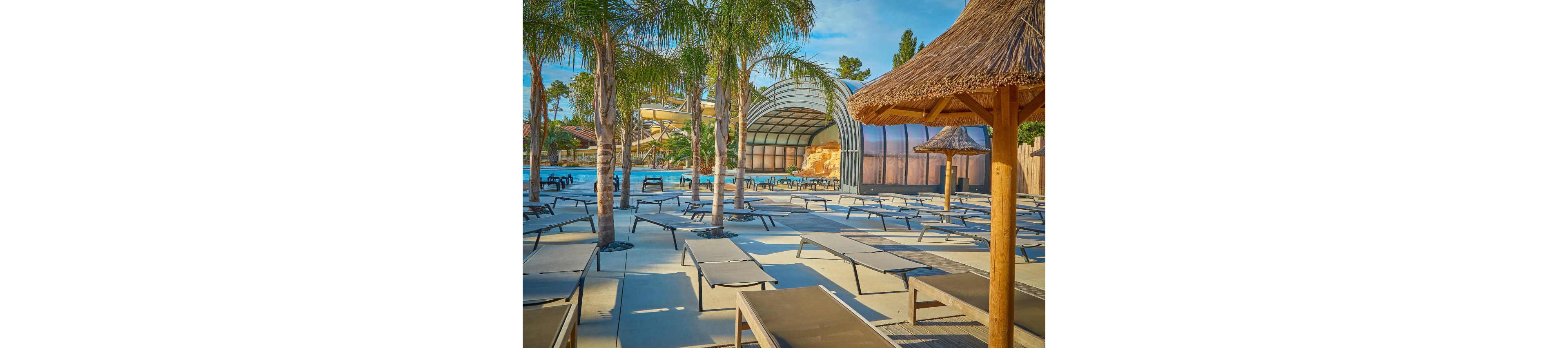 An outdoor resort pool area is lined with numerous empty sun loungers under tall palm trees. A covered pool structure and water slide are visible in the background, suggesting a family-friendly aquatic park. Straw parasols and warm-toned decking create a tropical vacation atmosphere. The bright daylight and clear sky enhance the relaxed, summery mood of the scene.