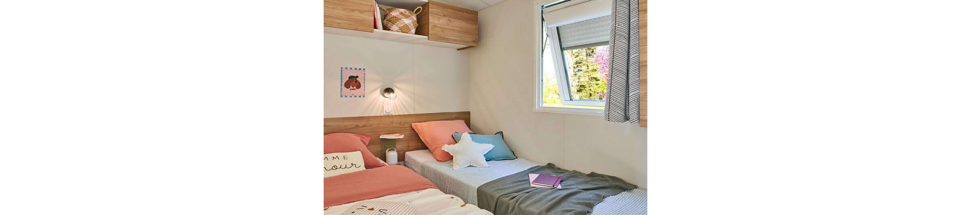 A small bedroom features two neatly made single beds with soft pastel bedding and decorative pillows. Wooden wall-mounted cabinets and a bedside shelf add storage and warmth to the space. Natural light enters through an open window, revealing greenery and pink blossoms outside. A wall lamp, framed artwork, and a notebook on the bed contribute to a relaxed, inviting atmosphere.