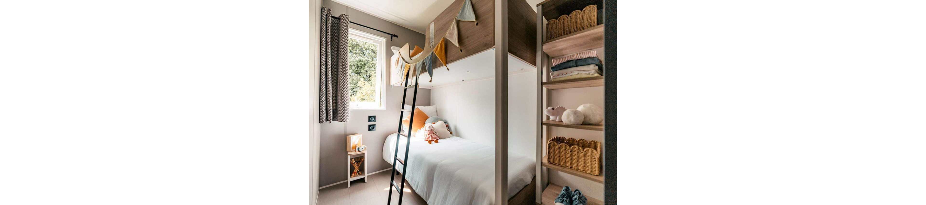 A small bedroom features a cozy bunk bed with a lower single bed neatly made with white bedding and accent pillows. A black ladder leads to the upper bunk, which is decorated with a colorful fabric pennant garland. Natural light enters through a window with patterned curtains, illuminating the neutral-toned walls. To the right, open shelving holds woven baskets, folded clothes, and plush toys, adding warmth and storage to the space.