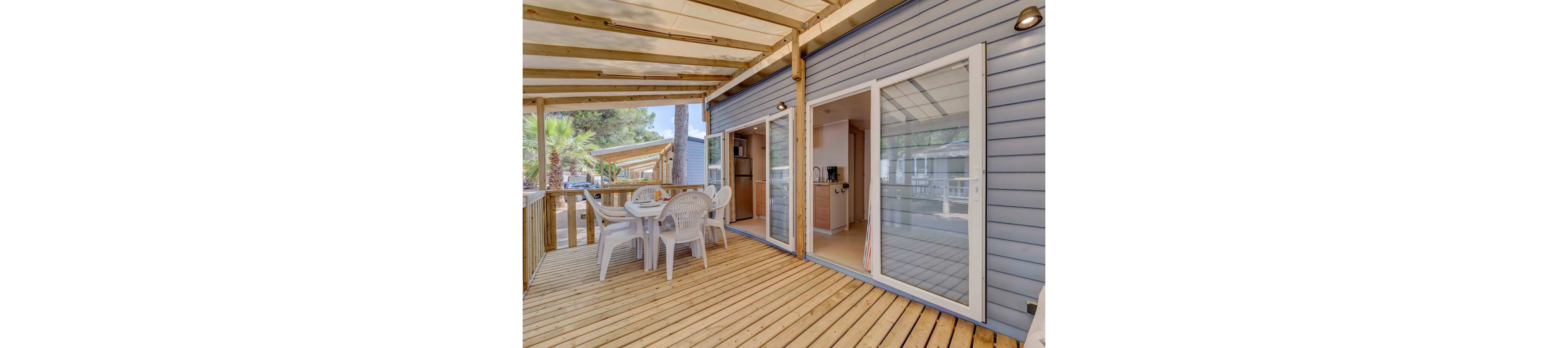 Image of a spacious wooden terrace attached to a modern mobile or modular home. The terrace features a white plastic dining table with several matching chairs arranged for outdoor meals. Large sliding glass doors open into a compact kitchen area with light-colored cabinetry and appliances. A wooden pergola roof provides shade, while surrounding trees and neighboring units suggest a holiday park or campsite setting.