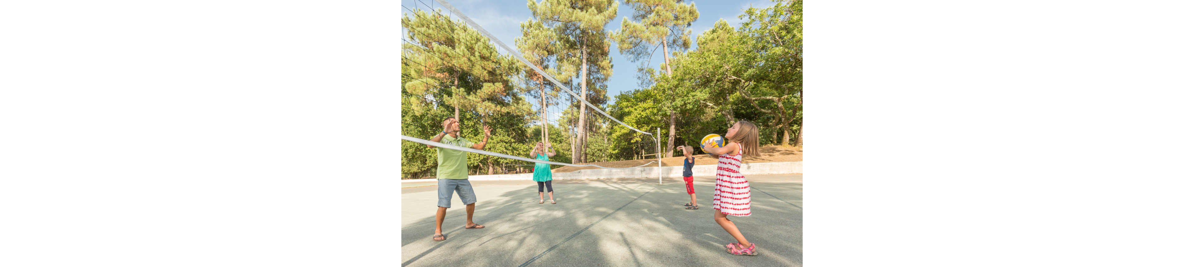 A family plays a casual game of volleyball on an outdoor court surrounded by tall trees. A young girl in a striped dress prepares to serve or pass the ball while holding it in both hands. An adult man and woman stand on the opposite side of the net, ready to receive, as a young boy watches nearby. The bright daylight and green forest backdrop create a cheerful, relaxed atmosphere.