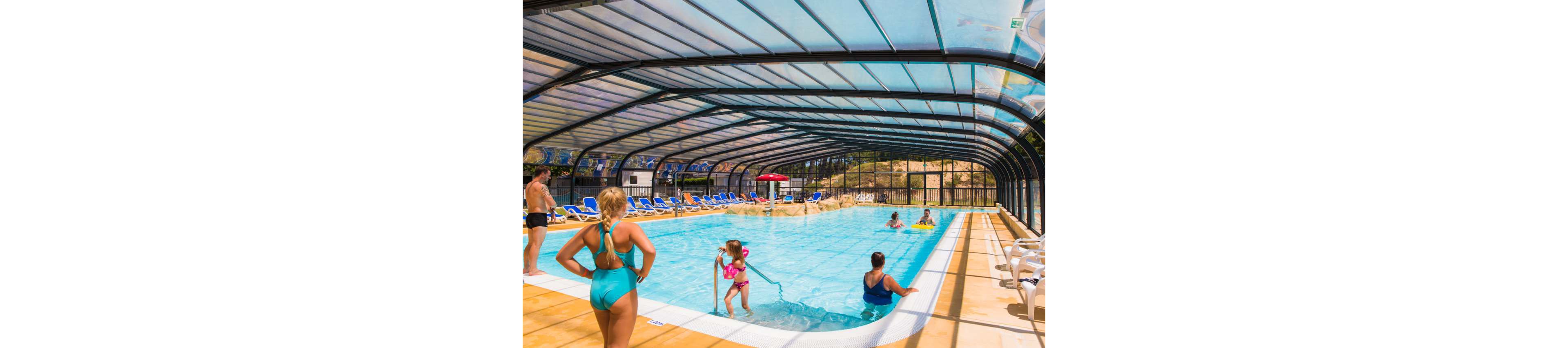 Several people relax and play in a large covered swimming pool. Adults and children are swimming, standing by the pool edge, and walking along the deck. Rows of blue lounge chairs line the sides, and the transparent arched roof lets in bright daylight. The scene suggests a family-friendly resort or campground pool area with a warm, leisurely atmosphere.
