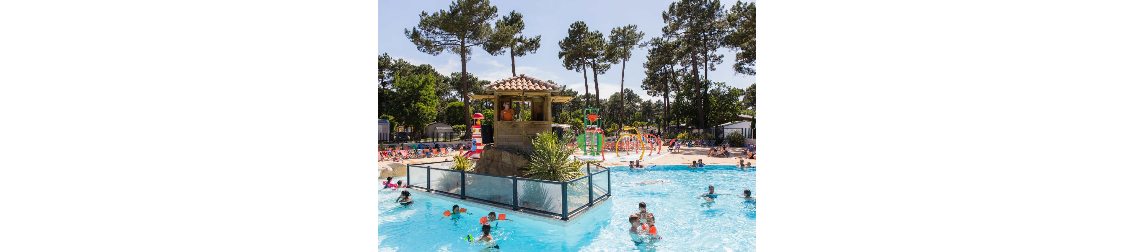 A lively outdoor swimming pool is filled with families and children playing in the water. The pool features a central wooden hut structure on a rock island, surrounded by glass safety barriers and tropical plants. In the background, colorful lounge chairs line the deck while people relax under the sun. A children’s splash area with bright water play structures and fountains adds to the fun atmosphere, all set among tall pine trees in a resort-like environment.
