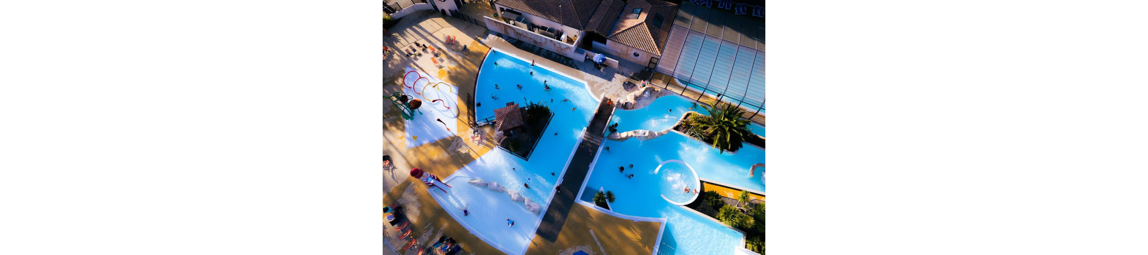 An aerial view shows multiple interconnected swimming pools at a resort. People of various ages are swimming, relaxing, and playing in the water. Surrounding the pools are lounge chairs, sunbathers, and a building complex with tiled roofs. Bright blue water contrasts with the warm tones of the deck and nearby greenery, creating a vibrant vacation atmosphere.