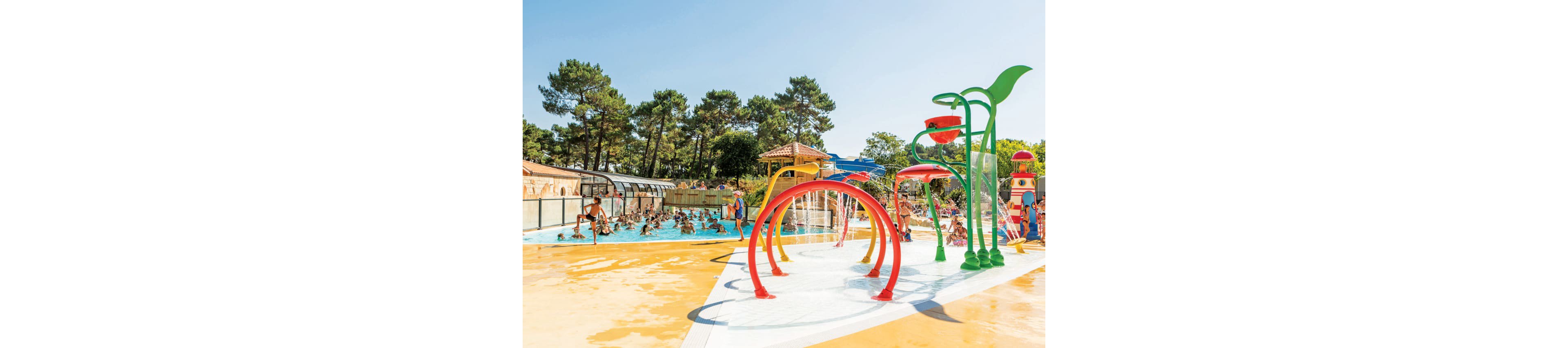 An outdoor family water park features a colorful splash area with curved spray arches and playful structures. Children and adults enjoy the shallow pools and water jets under a clear blue sky. A larger swimming pool in the background is filled with people, some under a partially covered area. Tall trees surround the park, creating a relaxed holiday atmosphere.
