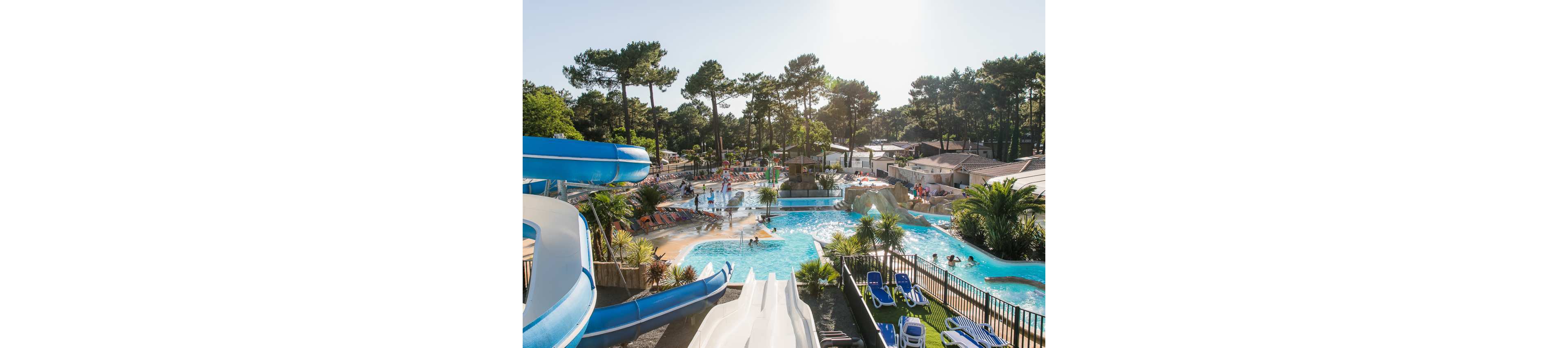 A lively outdoor water park features multiple pools surrounded by tall pine trees and holiday cabins. Large blue and white water slides curve into the main pool where people are swimming and playing. Sun loungers and umbrellas line the poolside, with families relaxing and children enjoying the attractions. The bright daylight and clear sky create a cheerful vacation atmosphere.