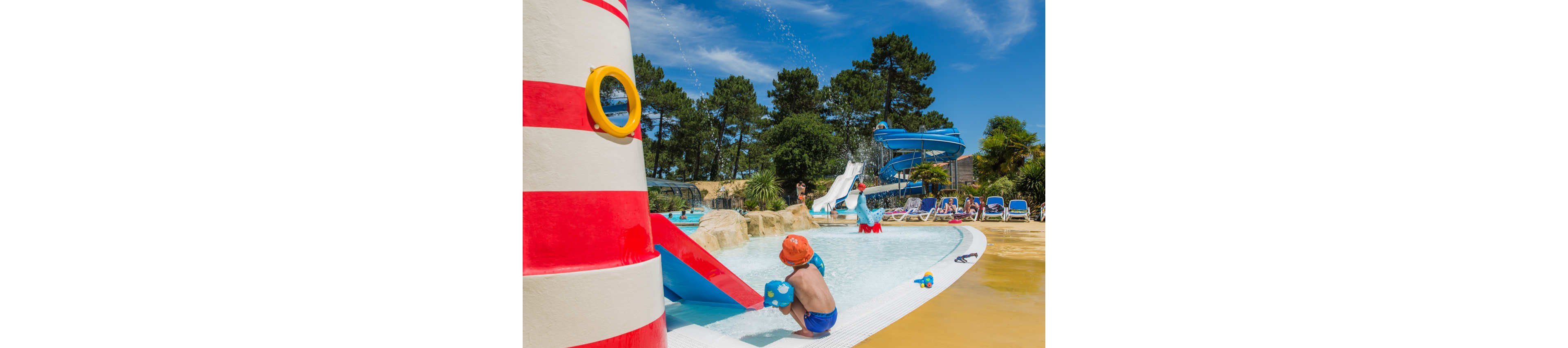 A young child in an orange hat plays at the edge of a shallow splash pool in a colorful outdoor water park. A striped lighthouse-themed structure sprays water while other children enjoy slides and water features in the background. Blue lounge chairs line the poolside where adults relax under a bright blue sky. Tall trees surround the area, creating a fun and family-friendly holiday atmosphere.