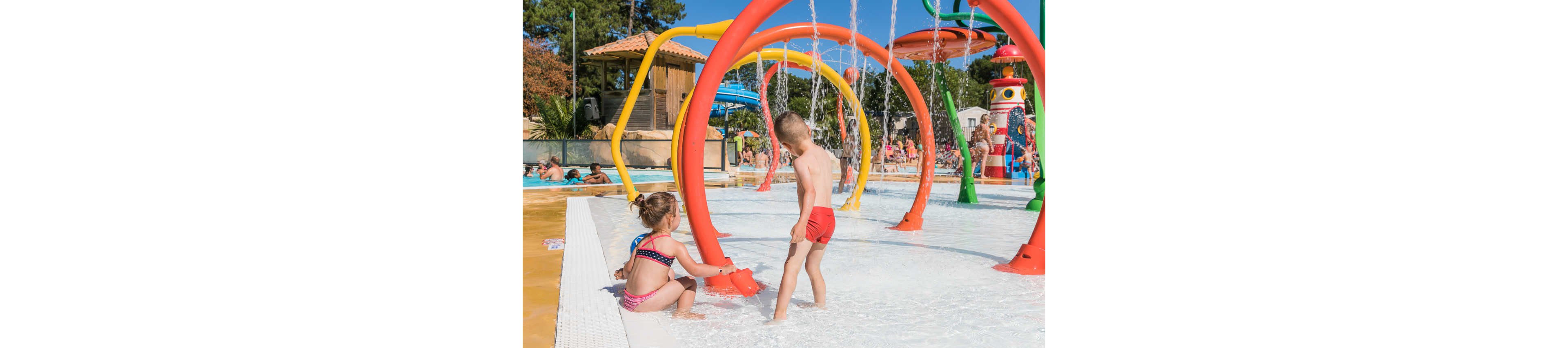 Two young children play in a shallow splash park surrounded by colorful water arches and fountains. The boy stands under a spray of water while the girl crouches near a red water feature. Bright primary colors and clear blue sky create a cheerful, summery atmosphere. Trees, buildings, and other park structures are visible in the background, suggesting a family-friendly outdoor recreation area.