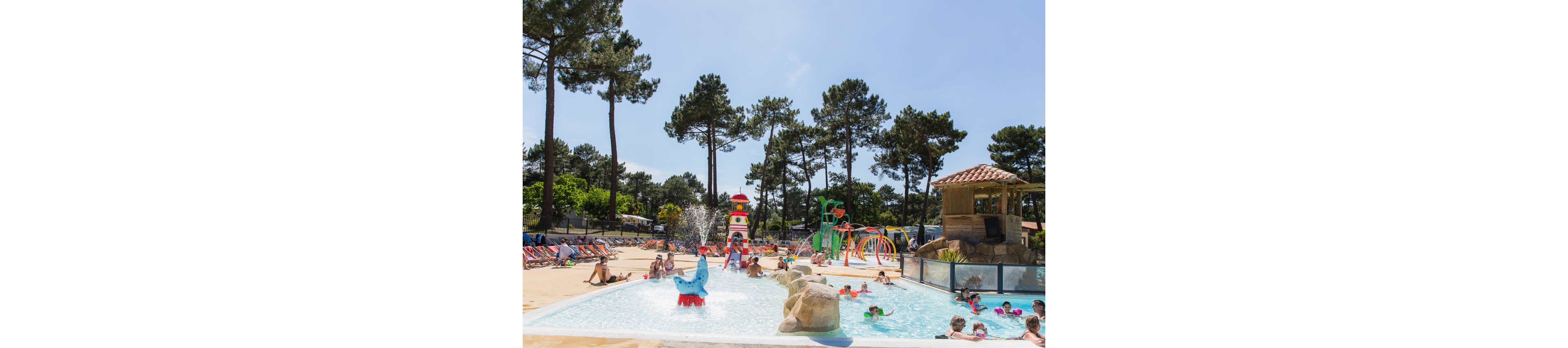 A lively outdoor water park features families and children playing in a shallow pool. Colorful slides, fountains, and splash structures create a fun and dynamic scene. Tall pine trees surround the area, providing a natural backdrop under a bright blue sky. Sun loungers and umbrellas line the poolside, suggesting a relaxed holiday atmosphere at a resort or campground.