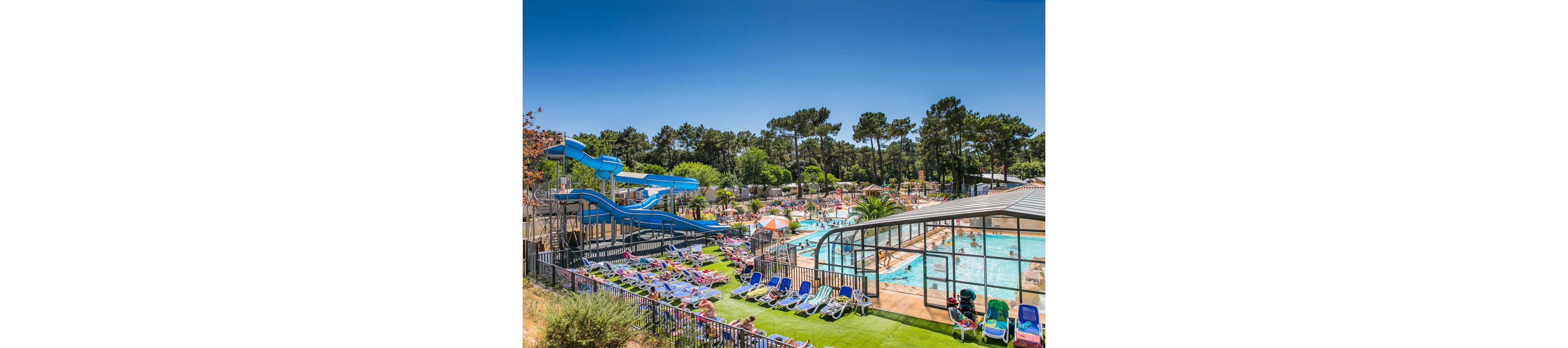 A lively outdoor water park features multiple swimming pools and bright blue water slides surrounded by tall trees. Numerous people relax on colorful lounge chairs and enjoy the sunny weather. A partially enclosed glass-roof pool area is visible on the right, adding a modern touch to the scene. The overall atmosphere is vibrant, family-friendly, and summery.
