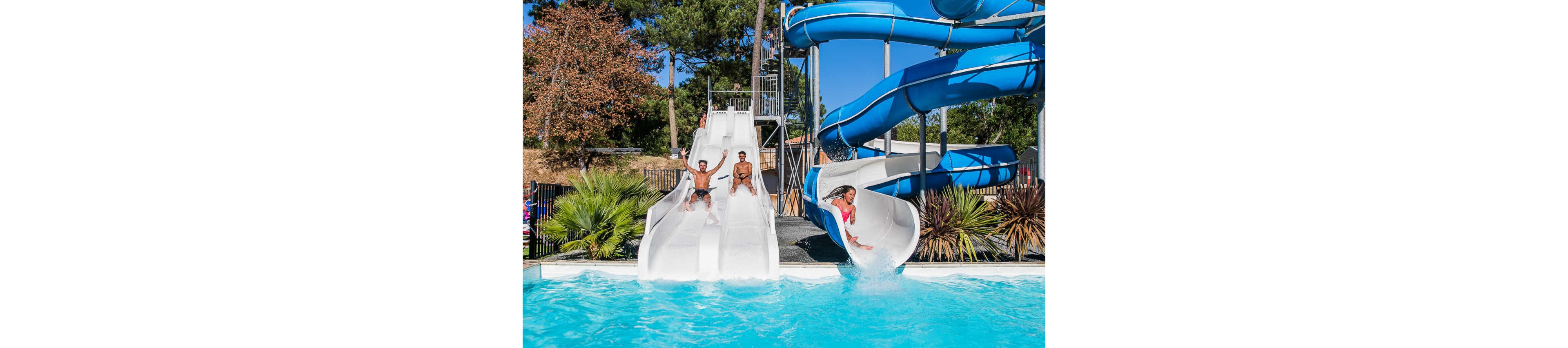 A group of friends rides down multiple water slides into a bright blue swimming pool. Two people slide side by side on white slides while another person splashes out of a curved blue slide. The scene is outdoors on a sunny day, surrounded by trees, shrubs, and a fenced area. The image captures energetic summer fun and playful movement in the water.