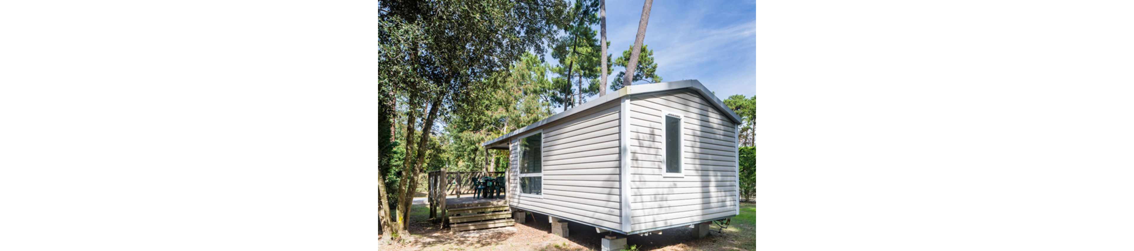 A light-colored mobile home cabin is set among tall trees in a wooded campsite. The structure is elevated slightly above the ground on supports and features a large window and a narrow vertical window. A wooden deck with outdoor chairs is attached to the side, suggesting a space for relaxation or dining. Sunlight filters through the trees, creating a calm and natural atmosphere around the cabin.