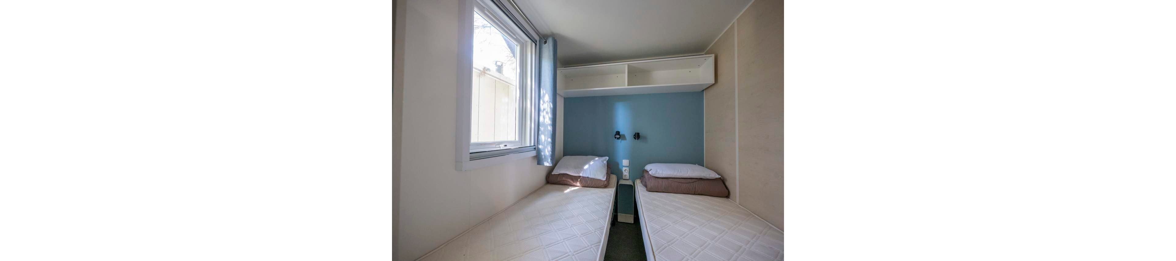 A compact bedroom features two single beds separated by a small nightstand. The room has a large window with a light curtain allowing natural light to brighten the space. A blue accent wall behind the beds includes two mounted reading lights and electrical outlets. Open shelves above the beds provide additional storage, creating a simple and functional sleeping area.
