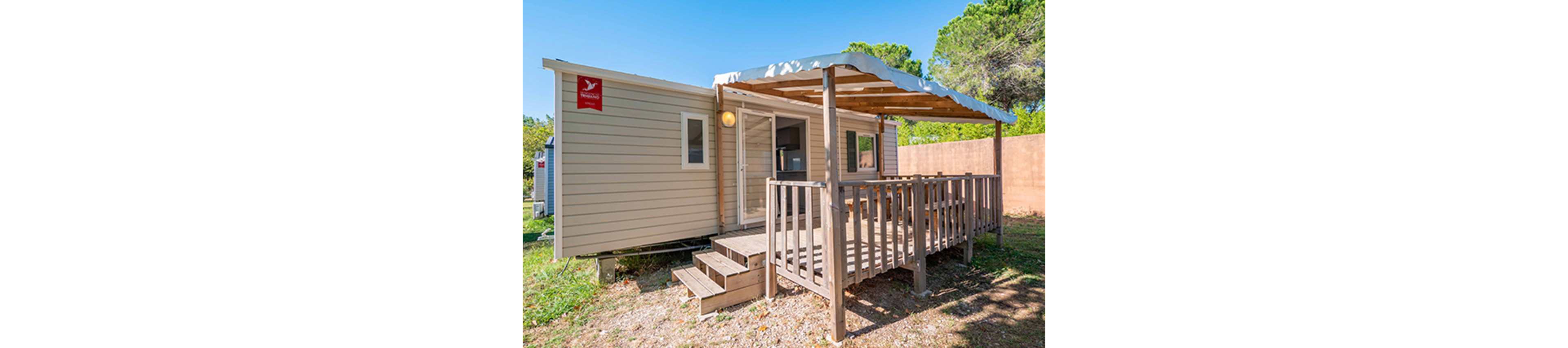 A beige mobile home with a small covered wooden deck is set in a sunny outdoor area. The structure features a front door, windows, and a short set of steps leading up to the porch. A wooden pergola provides shade over the deck, which includes a simple railing and outdoor seating. The home is surrounded by grass, trees, and a tall wall in the background, suggesting a quiet campsite or vacation park.