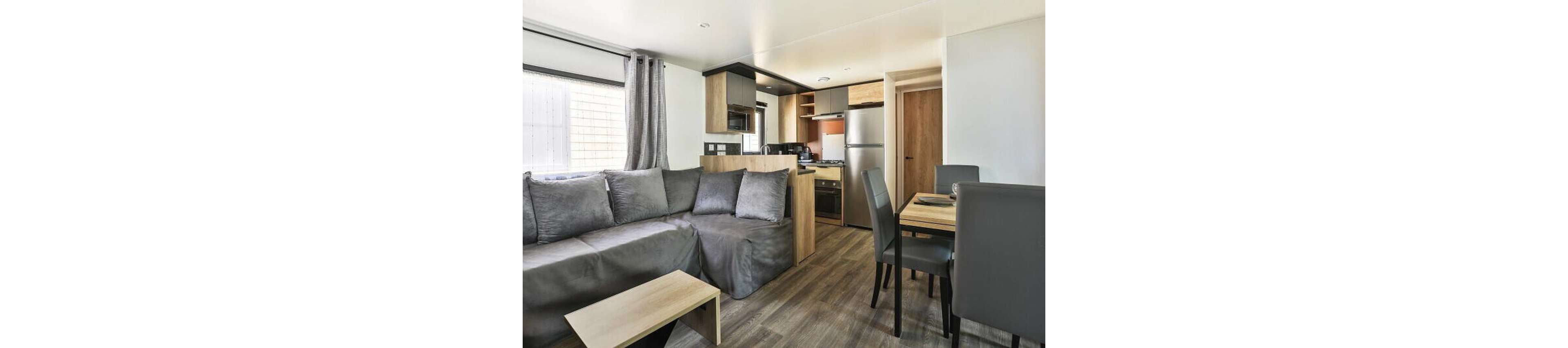A modern open-plan living space features a gray sectional sofa with matching cushions. The room includes a small wooden coffee table and a dining area with a wooden table and dark upholstered chairs. In the background, a compact kitchen with wood and black cabinetry, a stainless steel refrigerator, and built-in appliances is visible. Large windows with curtains allow natural light to brighten the neutral-toned interior.