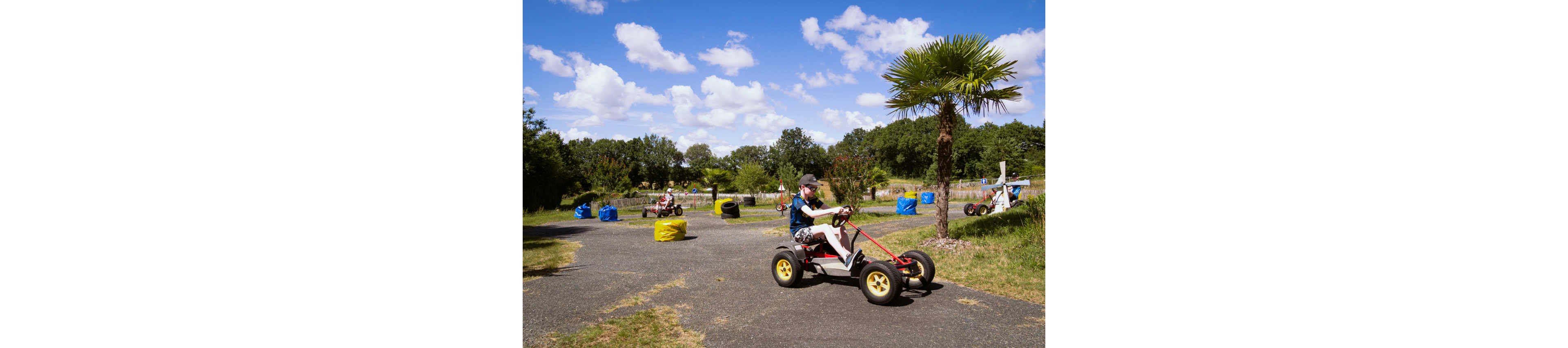 A child rides a pedal go kart on an outdoor track under a bright blue sky with scattered clouds. Other children drive similar karts in the background, navigating around colorful padded barriers. The track is surrounded by green grass, trees, and a small palm tree, creating a relaxed park-like setting. The scene feels playful and energetic, capturing a fun recreational activity for kids.