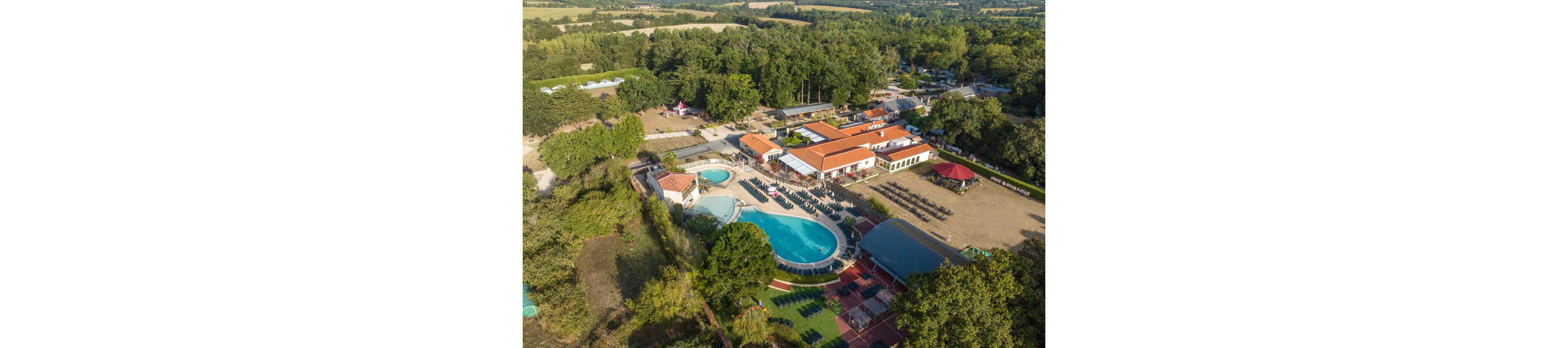 A large countryside resort complex is captured from above, featuring multiple swimming pools and sun loungers. The central buildings have red-tiled roofs surrounded by trees and open fields. Guests can be seen around the pool area and on nearby terraces, suggesting a relaxed holiday atmosphere. Additional facilities such as sports courts and shaded seating areas are visible within the property. The surrounding rural landscape emphasizes the secluded and tranquil setting.