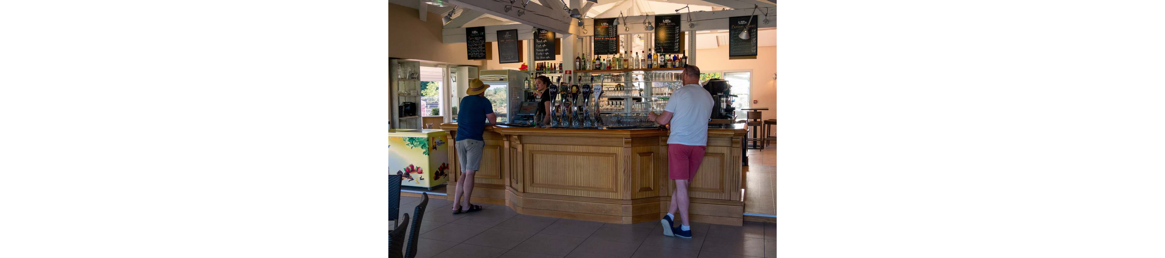 A bartender serves two customers standing at a polished wooden bar counter. The bar interior features glass shelves filled with bottles and multiple beer taps lined up on the counter. Chalkboard menus hang above the bar, and natural light enters from large openings, giving the space an airy feel. One customer wears a straw hat and casual clothes, while the other stands in shorts and sneakers, creating a relaxed, informal atmosphere.