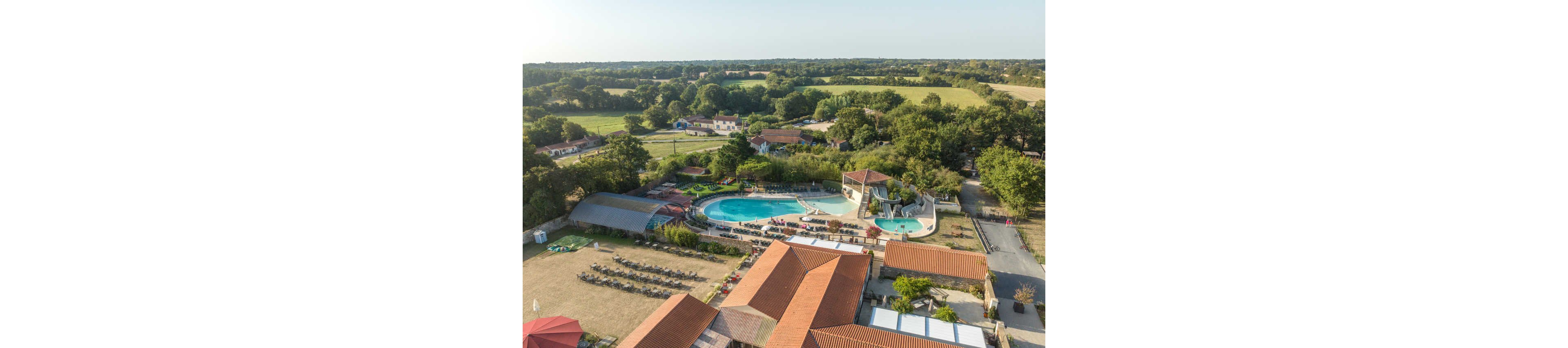 An aerial view shows a large outdoor swimming pool complex within a countryside resort. Curved pools, sun loungers, and landscaped greenery are surrounded by red-roofed buildings. A waterslide structure and a covered pool area add to the recreational facilities. Beyond the resort, open fields, scattered houses, and dense tree lines stretch into the distance under clear skies.