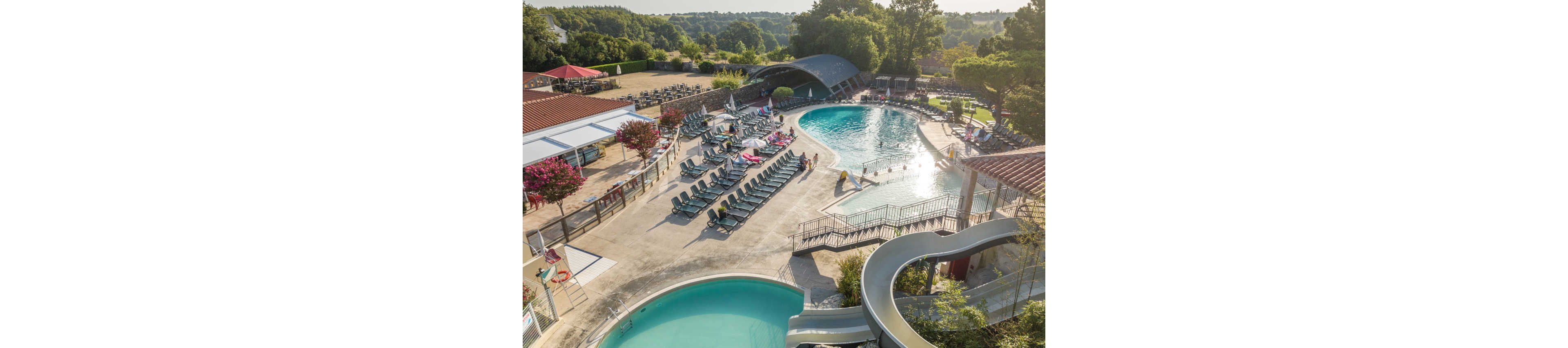 A large outdoor resort pool complex is captured from an elevated aerial perspective. Multiple curved swimming pools, including a main pool and a smaller circular pool, are surrounded by rows of sun loungers and parasols. A water slide winds down into one of the pools, and a few people can be seen relaxing and walking around the deck. The scene is set amid lush greenery and rolling hills in the background, suggesting a tranquil vacation setting. Bright sunlight and clear skies create a warm, inviting atmosphere.