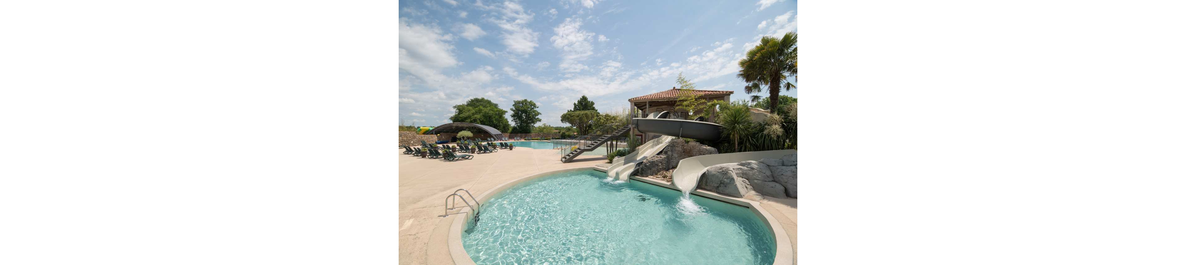 An outdoor swimming pool complex features a small round pool in the foreground with two curved water slides flowing into it. In the background, a larger pool area includes additional slides, lounge chairs, and shaded seating under a canopy. The scene is set on a sunny day with scattered clouds, palm trees, and landscaped greenery around the pools. The overall atmosphere suggests a relaxing vacation resort or family water park.