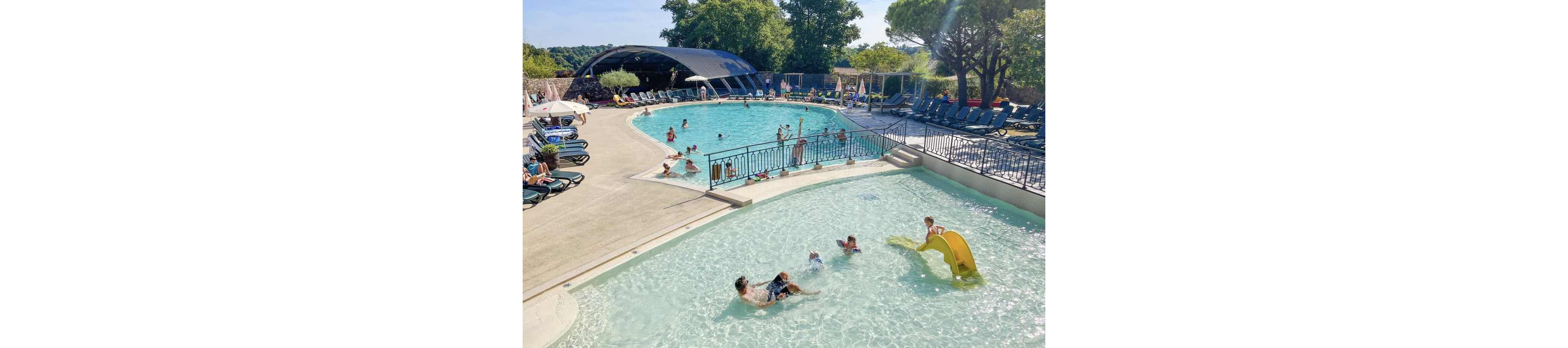 A large outdoor swimming pool complex is filled with families enjoying a sunny day. In the foreground, children and adults play in a shallow wading pool with a small yellow slide. The main pool curves around with people swimming and relaxing in the water. Numerous lounge chairs line the deck, and a covered area provides shade near the back. Trees and greenery surround the pool, creating a relaxed vacation atmosphere.